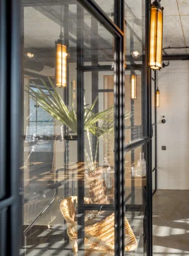 A modern, minimalist office space with large glass walls and black metal frames. The space is illuminated by long, industrial-style pendant lights. A potted palm tree is visible through one of the glass panels.